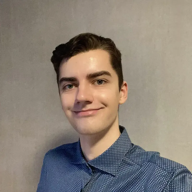 The person this portfolio is about, myself. I have short brown hair, wearing a blue and white patterned shirt, standing in front of a light grey plain wall, smiling at the camera.